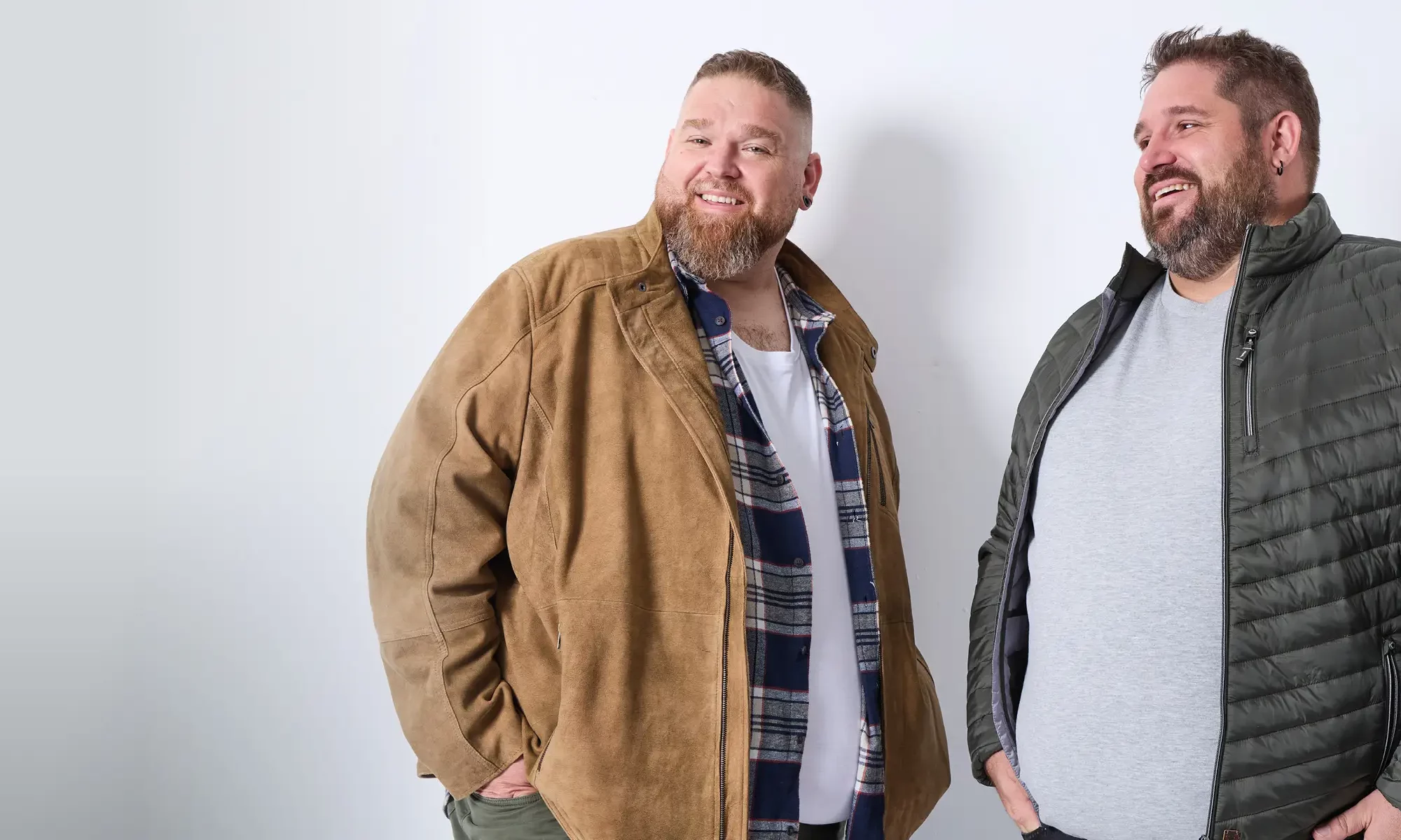 Zwei Männer mit Übergröße tragen stylische Outfits. Der linke in Karohemd und Lederjacke, der rechte in Steppjacke und T-Shirt.