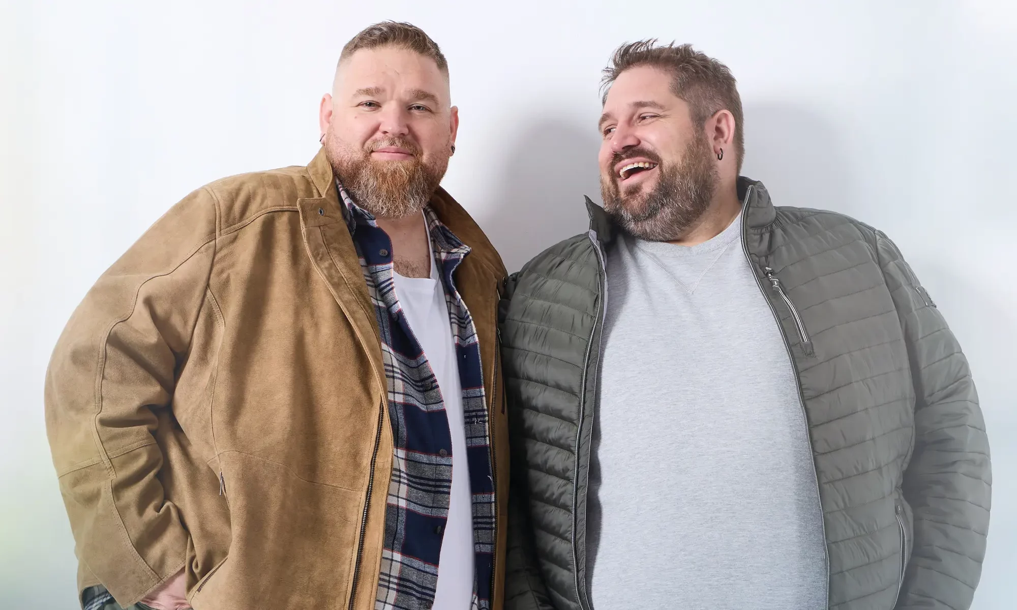 Zwei Männer mit Übergröße: links in Lederjacke mit offenem Karohemd und weißem T-Shirt, rechts in grüner Steppjacke mit grauem Pullover