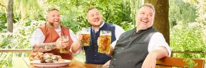 Drei Männer in Tracht trinken Bier und genießen eine Brotzeit im Biergarten in Freising.