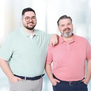 Zwei Männer mit Übergröße in eleganten Date-Outfits: Links ein Mann in mintfarbenem Poloshirt und beiger Stoffhose, rechts ein Mann in rotem Poloshirt und dunkelblauer Stoffhose.